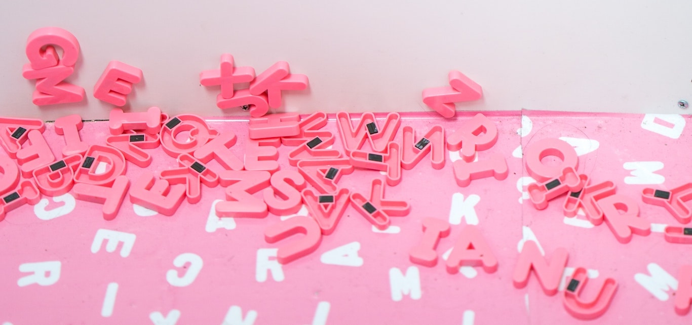 Kieli on merkitysten ja symbolien monimutkainen vyyhti. Vaaleanpunaisia kirjainmagneetteja levällään vaaleanpunaisella tasolla, jossa on valkoisella merkittynä valkoisella kullekin kirjaimelle oikea paikkansa.