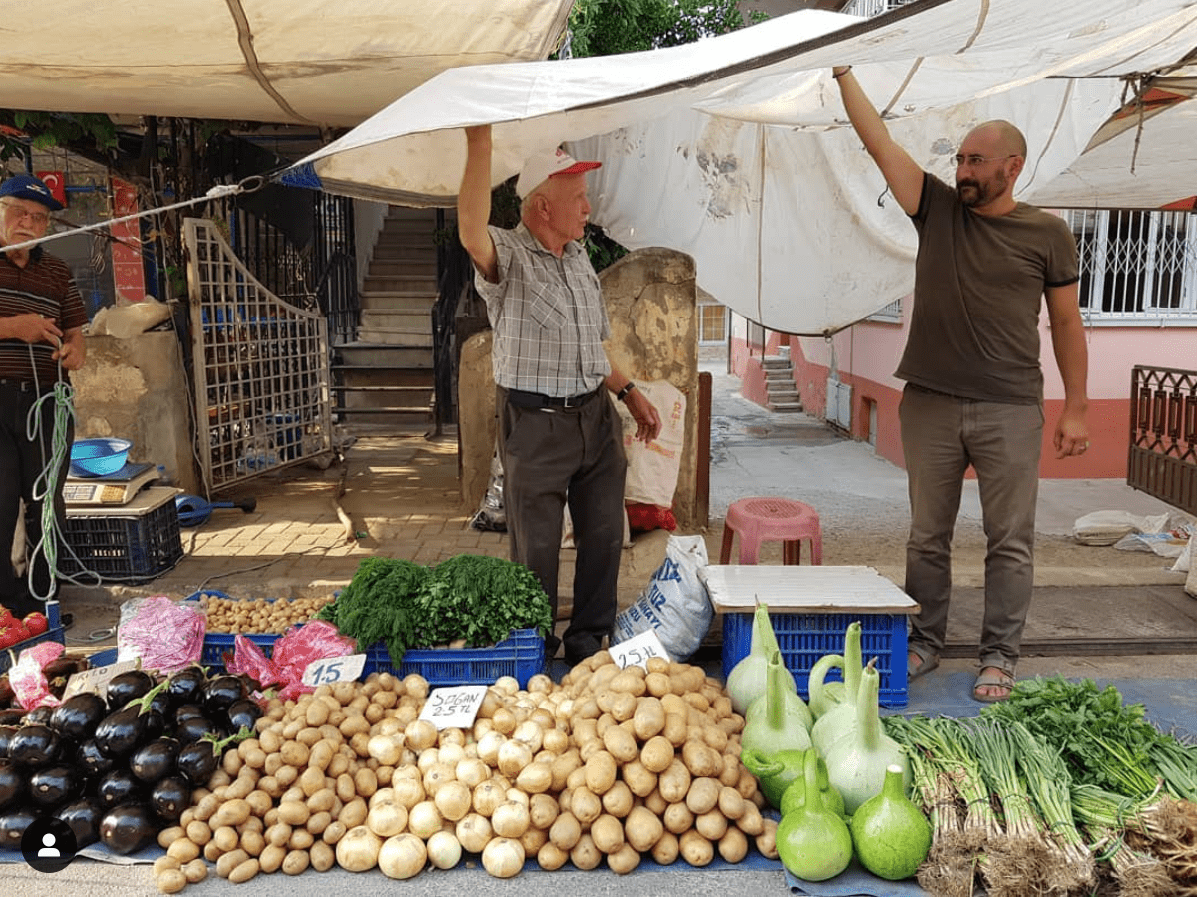 Tangör Tan seisoo lippalakkipäisen ruoantuottajan kanssa valkoisen pressun alla, joka on viritetty auringonvarjoksi. Etualalla torilla myytäviä vihanneksia.