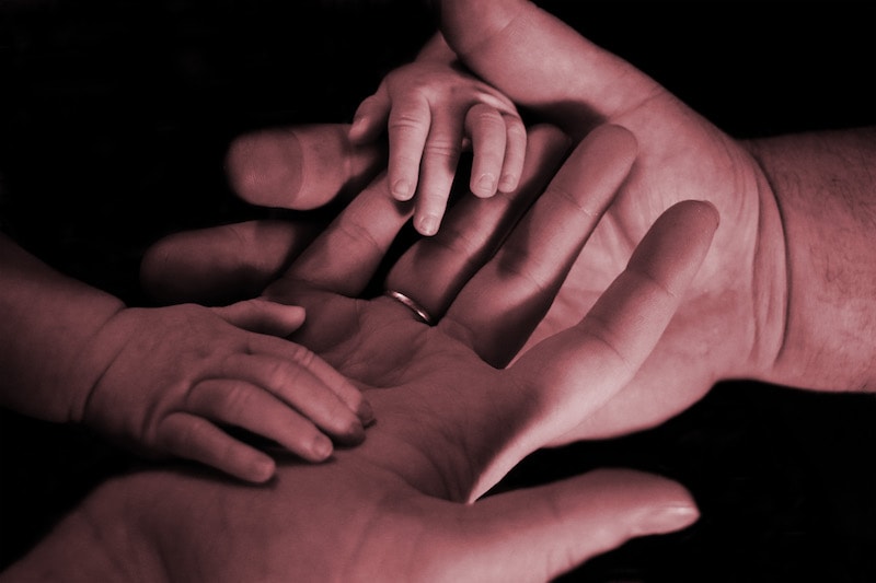 Vauvan kädet aikuisen käsien päällä. Aikuisen vasemmassa nimettömässä on sormus.