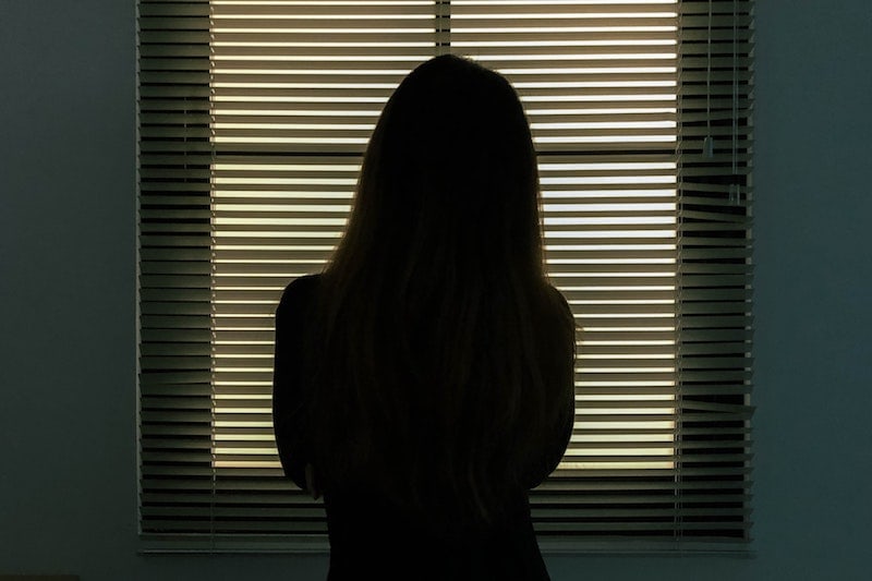 Vainoaminen aiheuttaa pelkoa. Nainen seisoo pimeässä huoneessa ikkunan edessä katsoen sälekaihdinten läpi ulos painostavan tunnelman vallitessa.