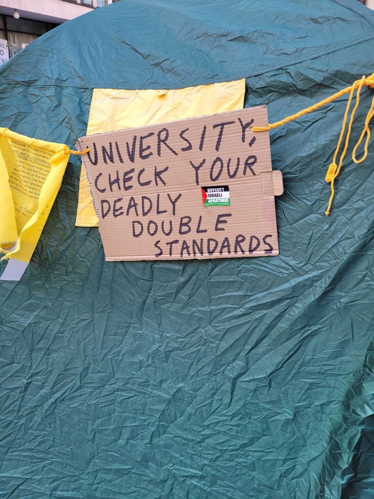 Hand-written sign with a text: University, check your deadly double standards.