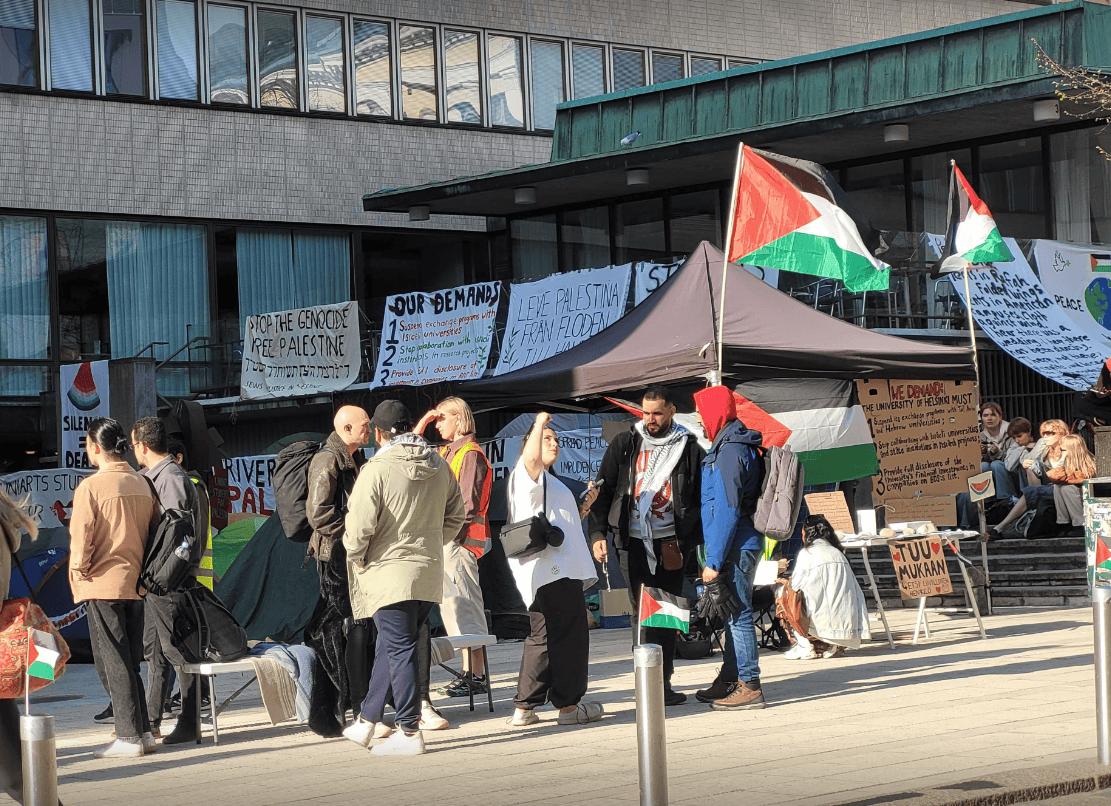 Students are standing and having conversations in front of Porthania building with hand written signs and flags of Palestine.
